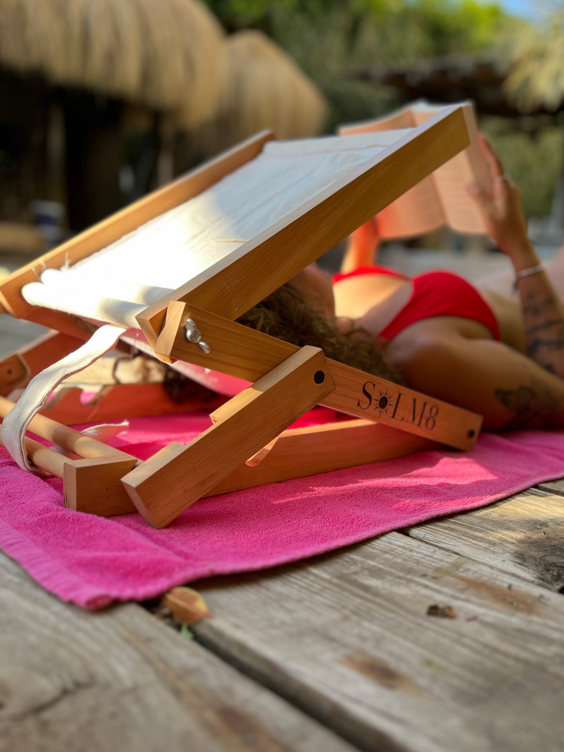 Person reading a book on a wooden deck with a pink towel and a wooden stand.